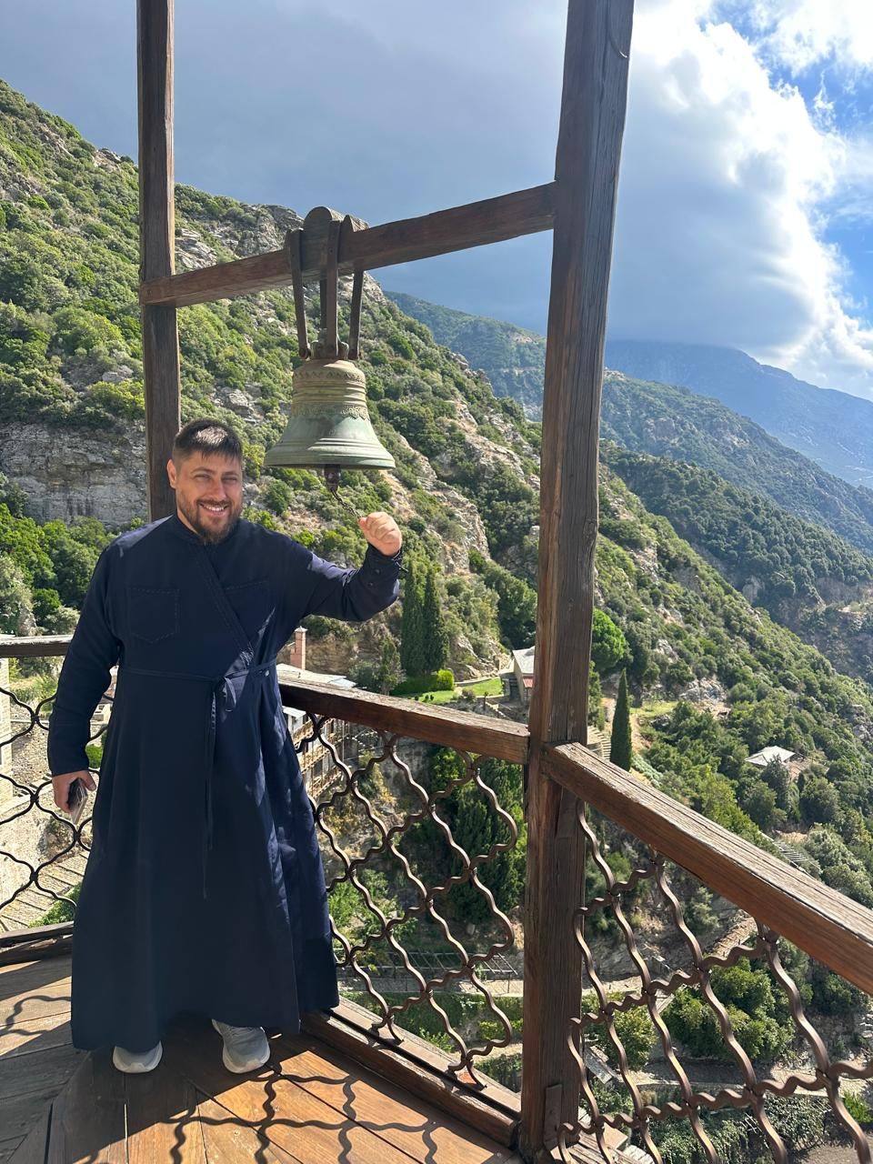 Священник Павел звонит в колокол афонского монастыря, за спиной горы